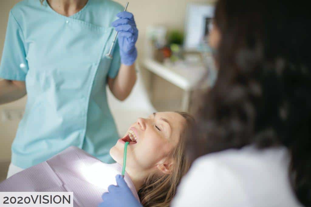 Crop faceless female dentist in uniform holding mouth mirror while preparing for inspecting female patient teeth with assistant holding tube suction in patient mouth